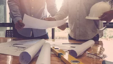 Two people reviewing building plans on a wooden table with rolled blueprints and a construction helmet.