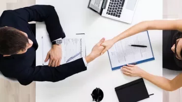 Two people shaking hands over a desk with documents and a laptop.