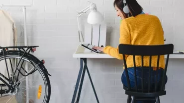 Person wearing headphones working on a tablet at a desk, with a bicycle and coat rack in the background.