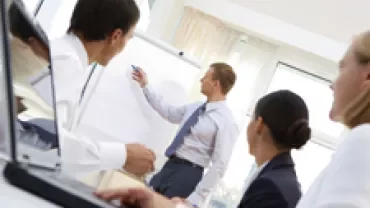 Group of people in a meeting, one person presenting at a whiteboard.