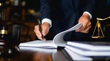 A person in a suit writes on a legal document at a desk with books and a gavel.