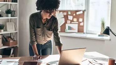 Person focused on a laptop at a desk with papers and graphs nearby.