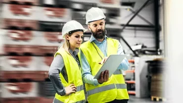 Two workers in safety gear review a tablet in a warehouse.