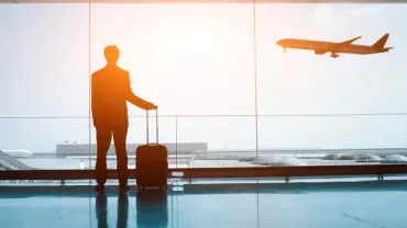 Person with suitcase watching airplane take off at sunset airport.