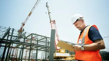 Construction site with a worker in a hard hat and safety vest writing on a clipboard.