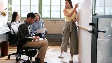 Group of people in a bright office, one writing on a whiteboard while others listen.