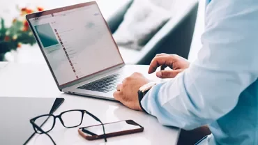 Person using a laptop on a white desk with glasses and a smartphone nearby.