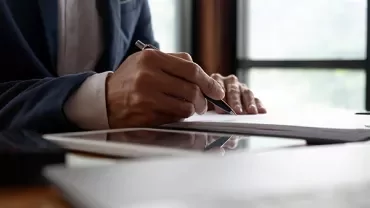 Person writing with a pen at a desk, near a tablet.