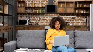 Person sitting on a gray couch using a laptop in a stylish living room with brick walls.