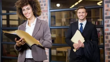 Two professionals in suits smiling, holding folders in an office setting.