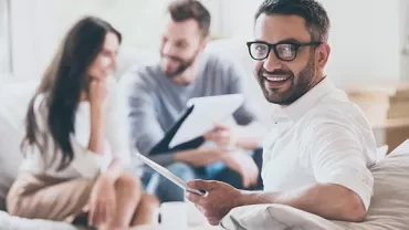 Smiling person with glasses holding a tablet, with two others on a sofa in the background.