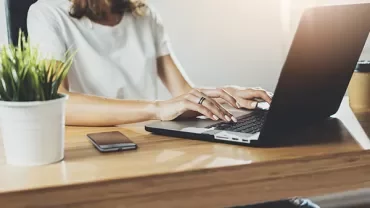 Person typing on a laptop at a wooden desk with a smartphone and plant nearby.