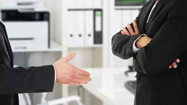 Two individuals in suits, one extending a hand for a handshake.