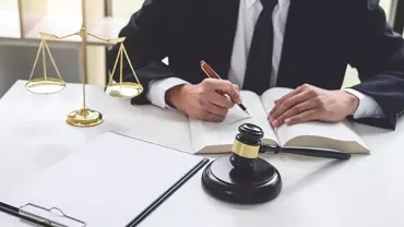 Person writing in a legal book beside a gavel and scales of justice.
